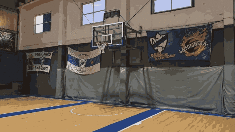 Vista interior de la cancha de básquet de Ferrocarril Midland. Se ve el piso de parqué, el aro con protecciones en la base y tres banderas azules y blancas colgadas en la pared gris del fondo, destacando una de "Mini Básquet" y otra con la frase "La Milineta"